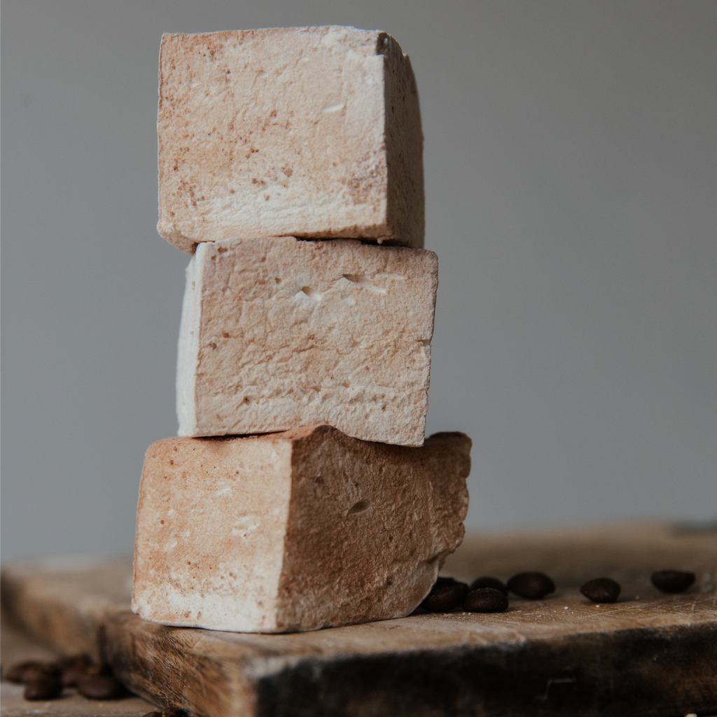 Stack of three beige-colored marshmallows on a wooden surface with coffee beans.